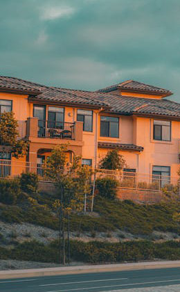 A large, modern two-story house with a sloped roof, surrounded by greenery and a fence, under a cloudy sky.