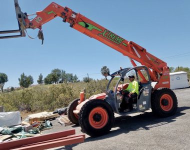 A bright orange telehandler is lifting materials on a construction site under a clear blue sky, surrounded by trees and equipment.