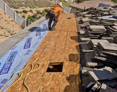 A roofer in an orange shirt works on a sloped roof covered with wooden boards and stacked tiles, preparing for repairs.