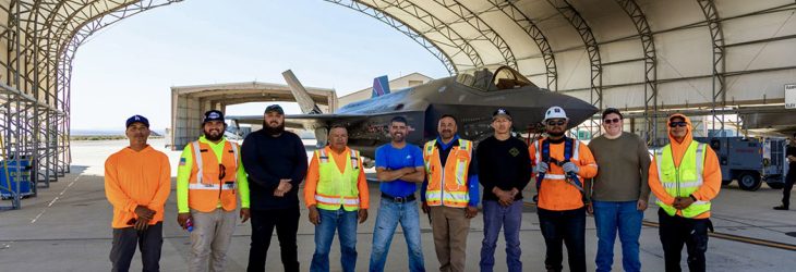 A group of men in orange vests stands in front of a fighter jet, showcasing teamwork and aviation expertise.