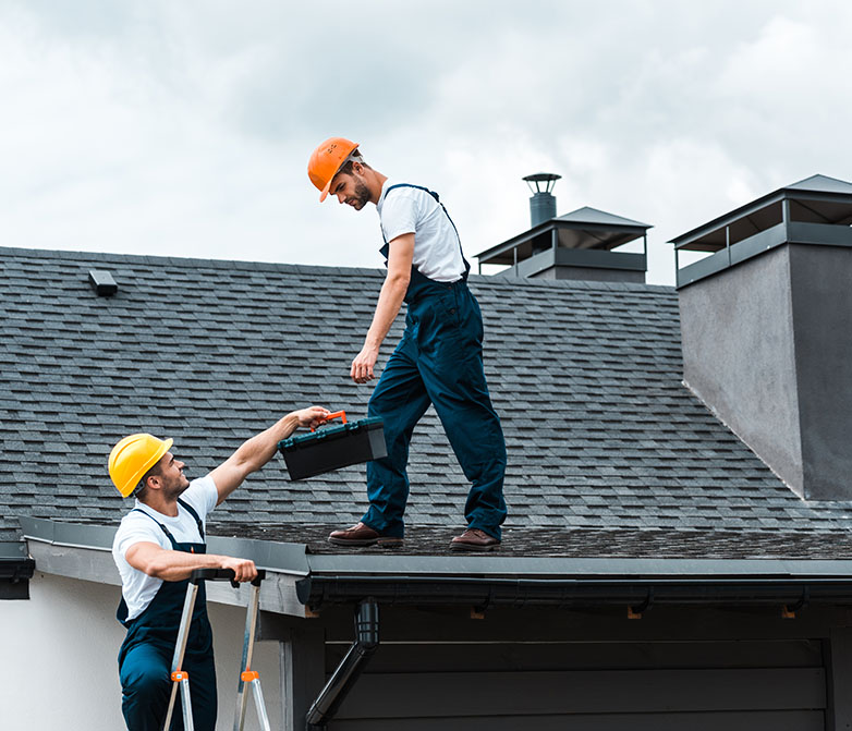 Two construction workers in helmets share tools on a rooftop, with one on a ladder handing a toolbox to the other.